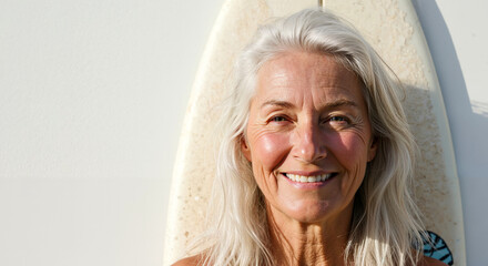 Senior female surfer smiling beside a surfboard against a wall  