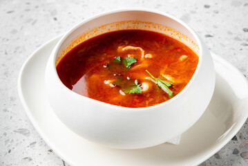 A bowl of spicy tomato soup garnished with fresh cilantro leaves, served on a white plate. The vibrant red color and herbs make the dish visually appealing and appetizing.
