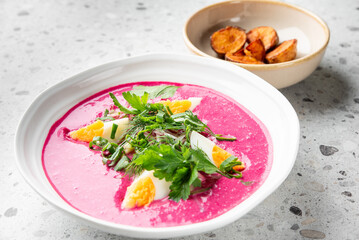 Vibrant pink beet soup with boiled egg quarters, fresh parsley, dill, and green onions in a white bowl. A side dish of roasted potatoes in a beige bowl on a speckled countertop.