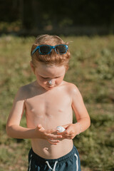 Little boy in swim trunks and sunglasses applying sunscreen on a sunny summer day. Outdoor scene...