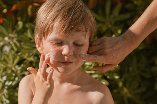 Mother&rsquo;s hands applying sunscreen to freckled face of young blond boy on a bright sunny day. Summer skincare, sun protection, family care and outdoor parenting concept.