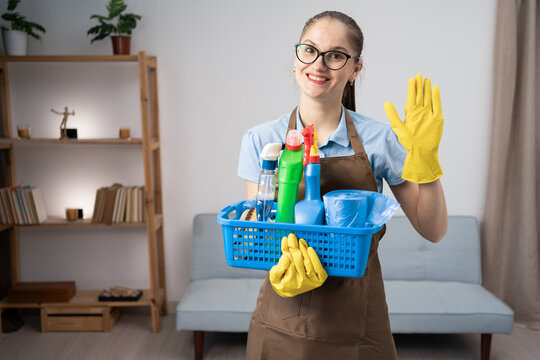 Female janitor with friendly welcome gesture. Brunette woman cleaner wearing apron holding cleaning products waiving hand and says hello