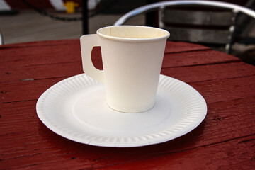 A white paper coffee cup with a handle placed on a white paper plate set on a rustic red wooden table in an outdoor cafe in Hamina, Finland
