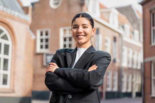 Confident young brunette realtor in sharp black suit posing with arms crossed outdoors, perfect for real estate marketing, career success, city professionalism, or personal branding