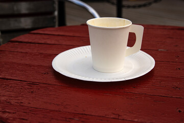 A white paper coffee cup with a handle placed on a white paper plate set on a rustic red wooden table in an outdoor cafe in Hamina, Finland
