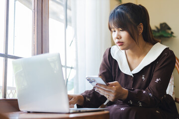 Asian woman working remotely as digital nomad uses smartphone and laptop in cozy home setting with natural light streaming
