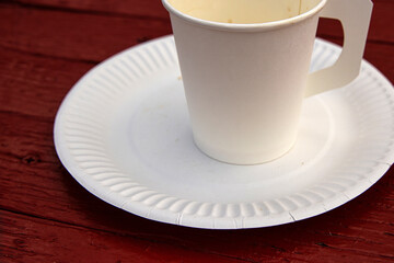A white paper coffee cup with a handle placed on a white paper plate set on a rustic red wooden table in an outdoor cafe in Hamina, Finland
