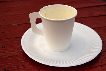 A white paper coffee cup with a handle placed on a white paper plate set on a rustic red wooden table in an outdoor cafe in Hamina, Finland