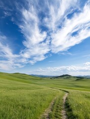 Obraz premium A winding dirt path cuts through a vibrant green field, leading towards rolling hills under a bright blue sky adorned with fluffy white clouds. The landscape evokes a sense of serenit : Generative AI