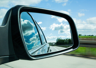Fototapeta premium Rear-view mirror showing an empty highway, cloudy blue sky, and green roadside — a symbolic image of road trips and freedom.
