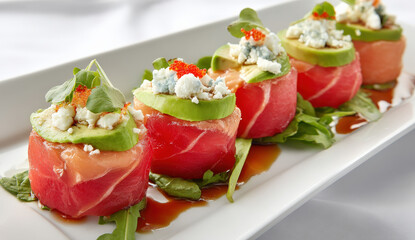 Four elegant tuna appetizers, each topped with avocado, blue cheese, and fish eggs, artfully arranged on a white rectangular plate with fresh greens and a dark sauce drizzle.