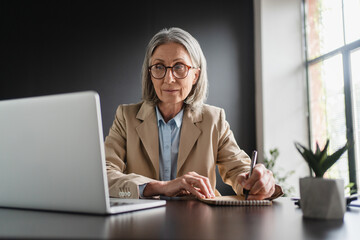 Busy concentrated mature business woman working in office using laptop writing notes. Focused 50...