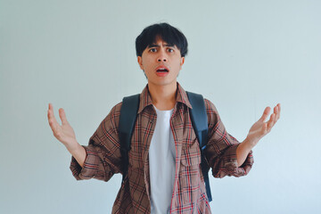Shocked male student wearing backpack looking at camera with confused expression, raising both hands, isolated on white background