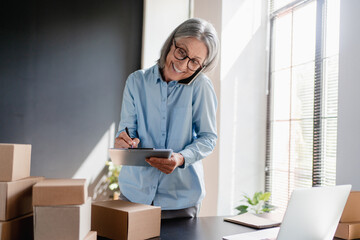 Inventory manager using smartphone packaging a parcel, preparing a small cardboard box for postage. Mature Caucasian small business owner working on laptop in warehouse. Dropshipping service.