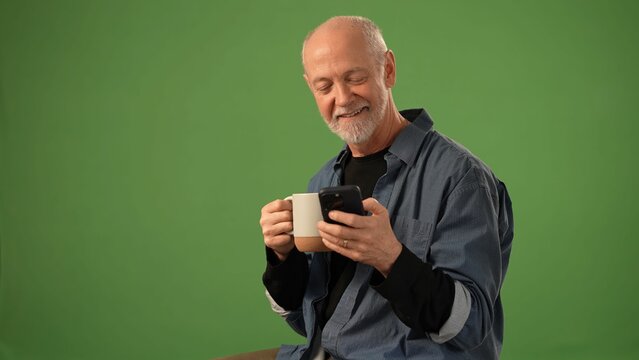Mature man smiles as he sips from a mug while viewing his smartphone, surrounded by a solid green background, ideal for announcements or product presentation. - Powered by Adobe