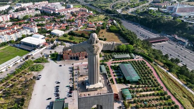 Almada, Portugal - May 12, 2024: Aerial shot of the Cristo Rei monument with arms outstretched, facing a dense residential cityscape and football field in Almada