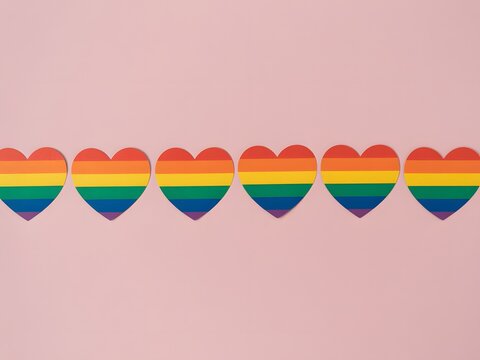 A flat lay top view of LGBT pride items: a rainbow colored paper hearts, placed on a pastel pink backdrop in line composition