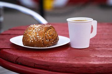 Takeaway coffee and pastry on cafe table. White paper cup and plate with cinnamon roll pastry  on...