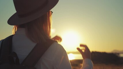 Traveler silhouetted against sunset, sun hat and backpack, forming heart shape with hands near serene lake horizon .Lifestyle, vacation, tourism, nature, active life - Powered by Adobe