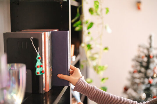 A person is selecting a book from a shelf. The books are on a dark shelf with a Christmas tree decoration nearby. The background is blurred.