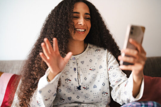 An Arab woman with curly dark hair is video chatting on her phone. She is wearing pajamas and waving to the person on the other end of the call.