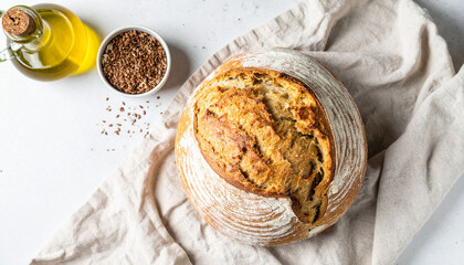 a rustic loaf rests upon a loosely draped cloth in this overhead shot. to the left, a clear bottle filled with golden liquid sits beside a small bowl of dark seeds