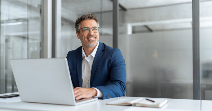 Middle age latin male entrepreneur businessman working on financial sale work at desk in office. Mature indian business man in suit using pc laptop for trading, looking smiling, dreaming at copy space