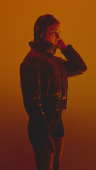 Side view portrait of young woman listening to music in headphones, wearing puffer jacket against orange background in neon lights. Concept of beauty and fashion, self-expression, human emotions. © master1305