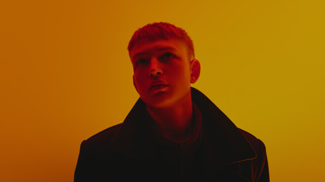 Portrait of stylish young guy in black coat and short hair style, looking upwards, posing against gradient yellow background in neon light. Bold fashion, style, youth culture, self-expression concept