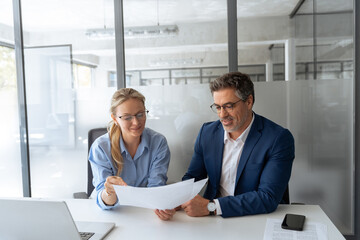 Team of mature businessman and accountant woman discussing financial tax document report sitting at table in office workplace. Two colleagues professional business people working together on project