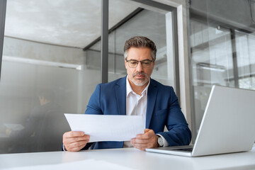 Busy latin mature business man working on laptop computer reading financial document report in...