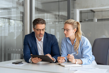 Team leader explain trainee working process using tablet pc computer. Two professional business people working together. Partners mature man and woman discussing project sitting at desk in office. 