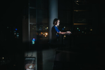 A dedicated woman sits at her laptop, illuminated by the screen's glow, working late into the...
