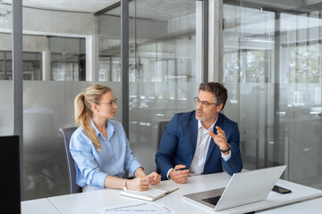 Two partners, colleagues, team of professional finance specialist business people working together. Mature mentor senior leader man, young woman manager discussing project sitting at desk in office