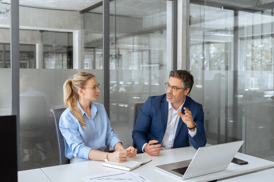 Two partners, colleagues, team of professional finance specialist business people working together. Mature mentor senior leader man, young woman manager discussing project sitting at desk in office - Powered by Adobe