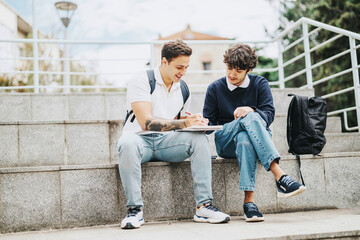 Two young students sit on outdoor steps, engaging in a study session with notes. They appear relaxed and focused, surrounded by an urban environment.