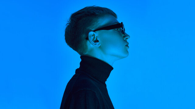 Side profile of stylish young man in turtleneck and sunglasses, posing under deep neon blue background ,expresses futuristic elegance. Concept of bold fashion, style, youth culture, self-expression