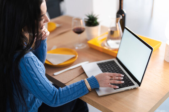 Woman works on her laptop at home. She is wearing a blue sweater and has a glass of wine nearby.
