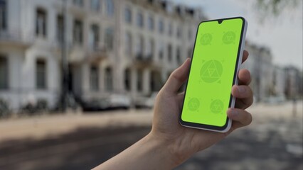 GREEN SCREEN CHROMA KEY person holding smartphone with green screen in sunny street with European architecture, filmed during daytime in Amsterdam