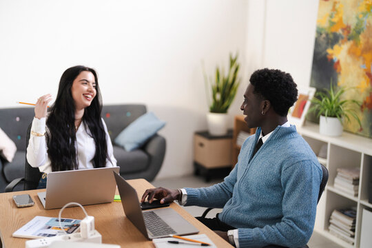 Two colleagues are working together at the office. They are using laptops and discussing a project, with one laughing at something the other said.