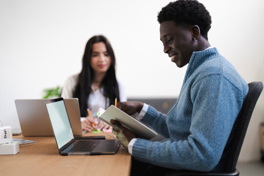 A young man reviews notes in a book at the office. A colleague works on a laptop in the background.