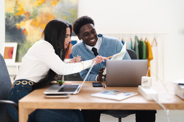 Two colleagues are collaborating on a project in their office. They are reviewing documents and using laptops to complete their work.