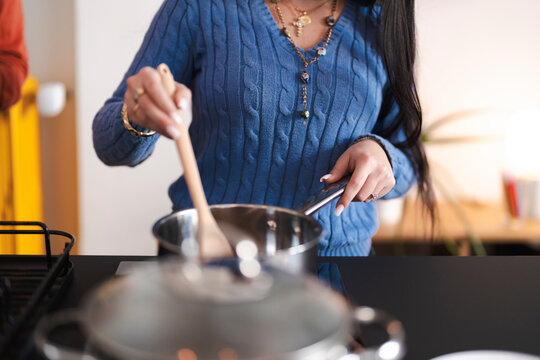 A woman in a blue sweater stirs food in a pot with a wooden spoon. She is cooking in a modern kitchen.