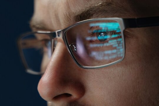 Close up of focused software engineer or data scientist wearing glasses, analyzing code on his screen. Programming script reflects on his eyeglasses as he works late.