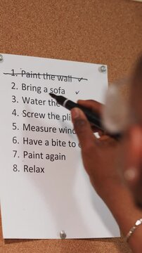 Black guy crosses out accomplished to-do list item on cork board near grunge wall closeup. African-American worker with plan of flat renovation process