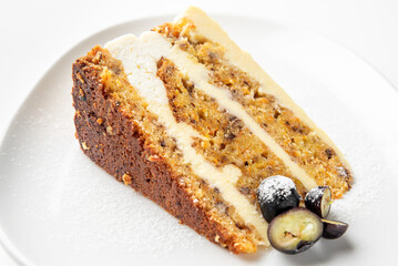 A close-up of a slice of carrot cake with cream cheese frosting, garnished with powdered sugar and sliced grapes, presented on a white plate. The cake's texture, moist layers, and rich flavors