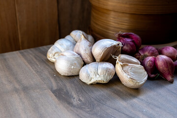 A collection of fresh garlic bulbs and red onions rests on a wooden surface, with a bamboo steamer visible in the background. Essential cooking ingredients.