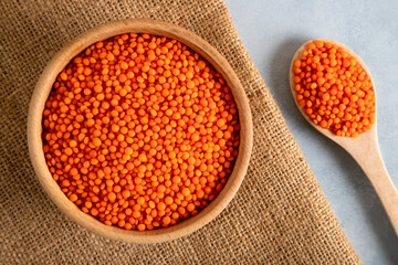 Red lentil grains in a wooden bowl and spoon on a light blue background. Top view of dried orange lentils inside the bowl.