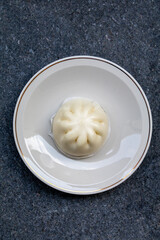 A single, perfectly steamed Baozi or steamed bun is served in a minimalist white bowl. Its soft, doughy texture and signature pleats are highlighted, emphasizing the simple elegance of this popular As