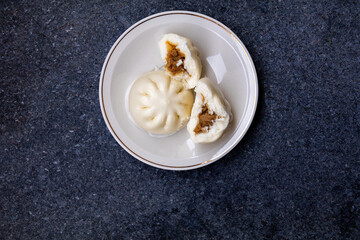 A top-down view of two steamed buns on a white plate with a gold rim, one whole and one split in...